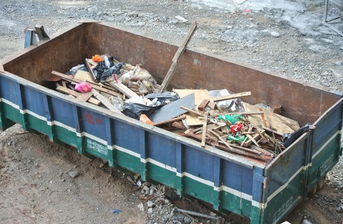 A rectangular metal skip with a rusted finish, positioned on an outdoor gravel surface, filled with a mixture of construction and garden waste. Visible materials include broken wooden planks, plastic bags, pieces of cardboard, and assorted debris, some of which are partially torn or crumpled. The skip is situated in a rough, unfinished area with loose gravel and broken concrete around its base. The scene suggests an ongoing waste clearance, typical of garden or property renovation projects in Forest Hill. The skip is partially filled and appears ready for rubbish removal by Garden Clearance Forest Hill, a company specializing in rubbish disposal and clearance services. Natural daylight illuminates the scene, casting soft shadows on the debris and emphasizing the variety of materials contained within the skip.