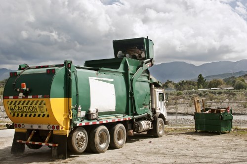 A large green industrial waste collection truck with a yellow caution stripe at the rear, parked on an unpaved area near a chain-link fence. The truck's rear compactor is lifted upward, revealing the open loading area. To the right of the truck, there is a small green waste container filled with miscellaneous rubbish, including some wooden planks and other debris. The ground appears to be a mixture of dirt and gravel, and the background features a cloudy sky with dark clouds and mountain ranges in the distance, suggesting an outdoor setting in a rural or semi-rural environment. The scene is typical of rubbish collection operations, aligning with the services offered by Garden Clearance Forest Hill for waste removal in the local area around Forest Hill, London postcode area.