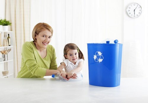 A woman and a young girl are seated at a white table in a bright, modern kitchen setting, both smiling and engaging in a recycling activity. The woman, wearing a light green top, has short blonde hair and is guiding the girl, who has dark hair tied in pigtails and is wearing a white shirt. They are working with a clear plastic bottle, preparing it for disposal or recycling. To the right of the image, a large blue recycling bin with a white recycling symbol is positioned on the table, indicating waste separation. The background features white blinds covering windows, a small clock on the white wall, and a few storage shelves with decorative items. The scene emphasizes eco-friendly waste management and household rubbish disposal, aligning with services offered by Garden Clearance Forest Hill in the local area, potentially serving residents in the Forest Hill post code district.
