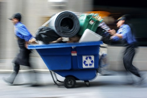 A blue wheeled waste bin overflowing with various rubbish items is being pushed by two individuals wearing blue tops and dark trousers, possibly city workers or clearance staff. The bin contains a large black plastic bag, a green plastic recycling bin, a white object, and other assorted waste materials. The background appears to be a street scene with a blurred vehicle passing by, indicating motion. The environment suggests a typical urban or suburban setting, likely associated with rubbish collection or clearance services commonly found in Forest Hill or surrounding areas. The image illustrates the process of rubbish removal, highlighting the practical aspect of waste disposal and collection efforts in the local community.