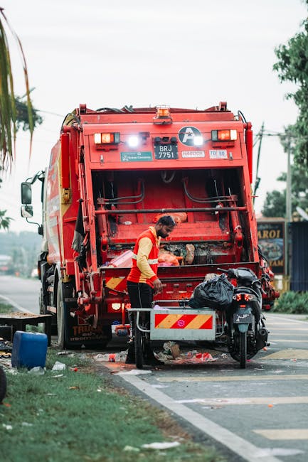 A street scene featuring a white refuse collection truck parked alongside a cobblestone pavement, with its rear hatch open revealing a rusty, metal interior designed for waste compaction. A worker dre