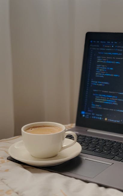 A silver laptop with an illuminated screen displaying lines of colorful computer code is placed on a white desk. To the left of the laptop, there is an open notepad with a pen resting on top. In front