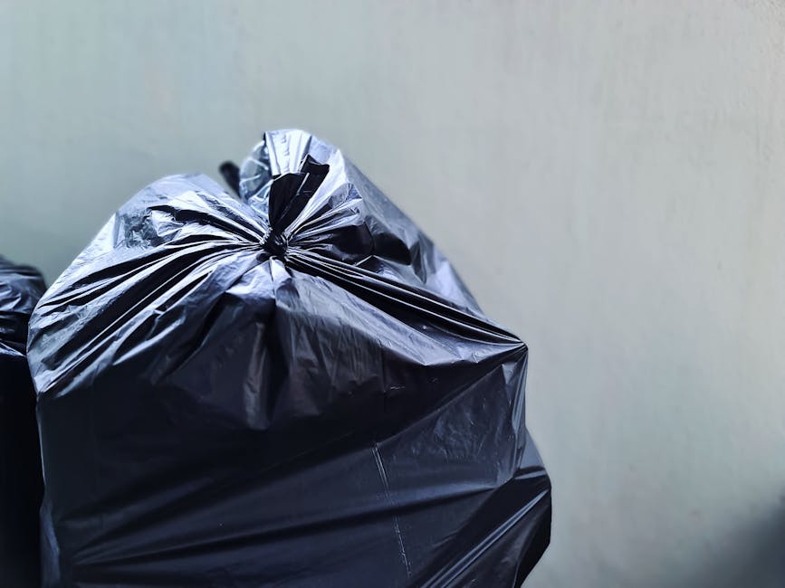 Close-up photograph of several large black garbage bags made of glossy plastic, tightly packed and sealed at the top with twisted closures. The bags are set against a plain, light-colored wall, possib